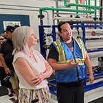 Tyrone Starr et la ministre Hajdu se tiennent devant 2 journalistes avec des caméras, lors de la visite de l'usine de traitement de l'eau de la Star Blanket Cree Nation.